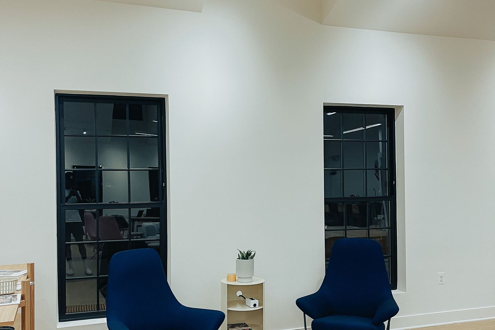 Counseling session room: two navy chairs facing each other beside a small table, black-framed windows looking into a commercial office